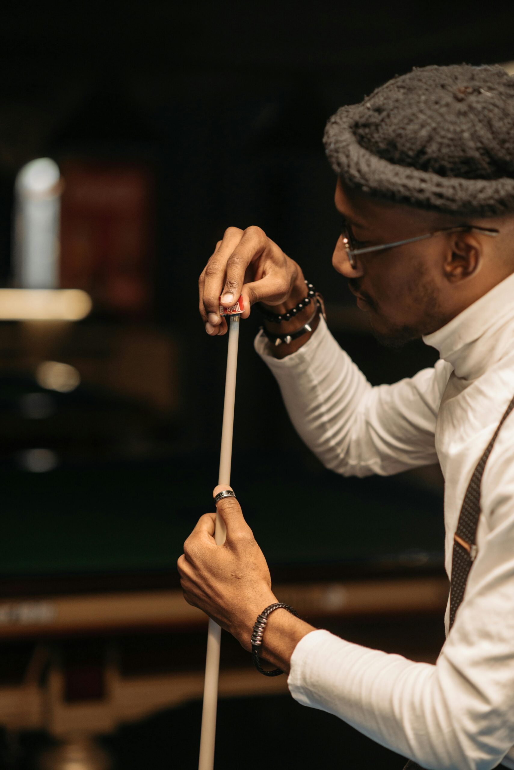A fashionable man sharpening a cue stick in an indoor billiard setting with a focus on style and precision.